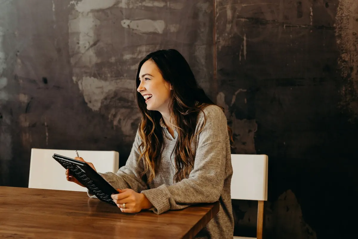 Une femme assise à une table, tenant une tablette et échangeant avec quelqu’un.